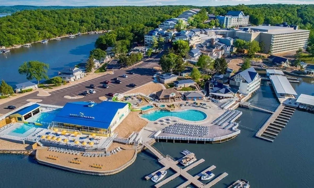 A lively exterior scene featuring a swimming pool with boats docked nearby, a beach area, and hotel buildings in the background. The setting is bustling with activity, evoking a sense of relaxation and fun around the water.