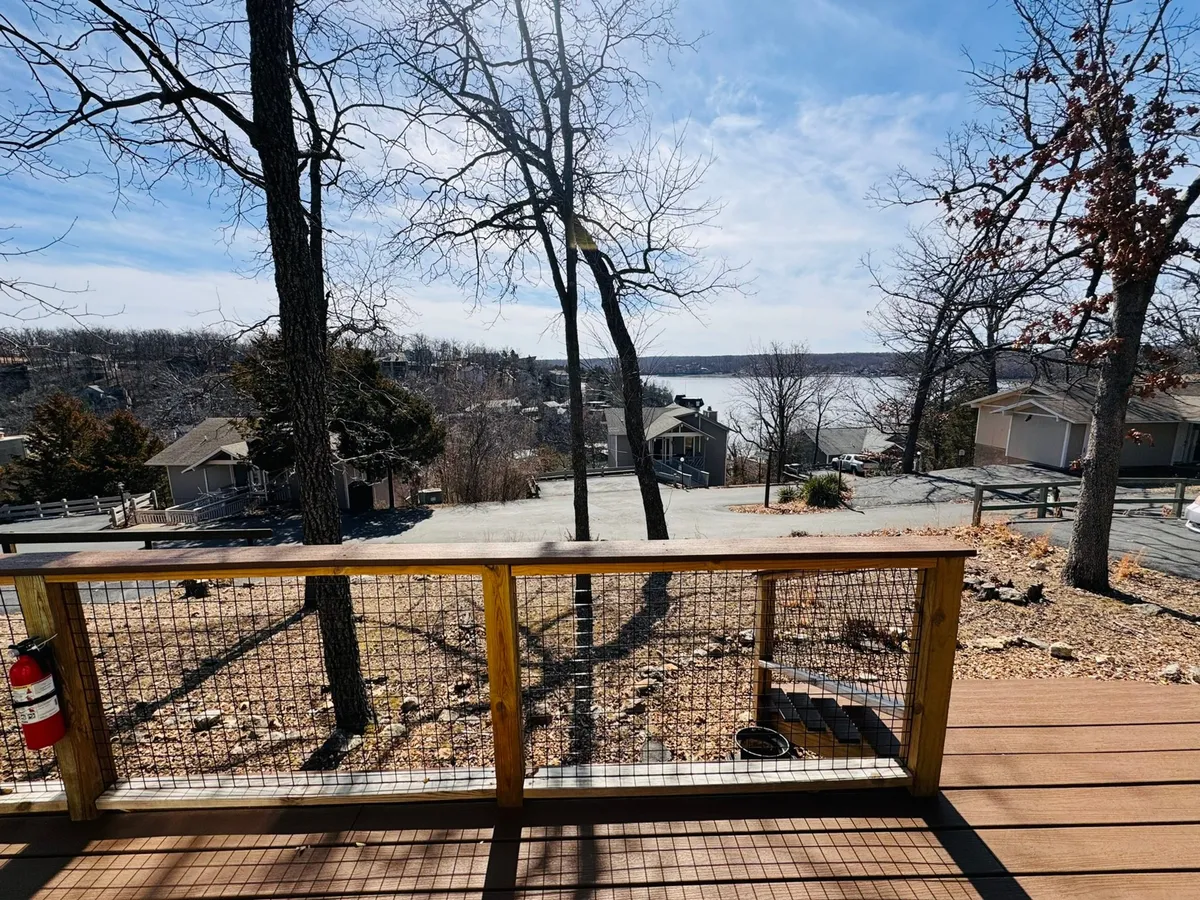 A tranquil lakeview setting with a deck railing in the foreground and lush trees surrounding the area. The deck appears spacious, ideal for group gatherings, with the serene lake providing a beautiful backdrop perfect for unwinding and socializing after a round of golf.