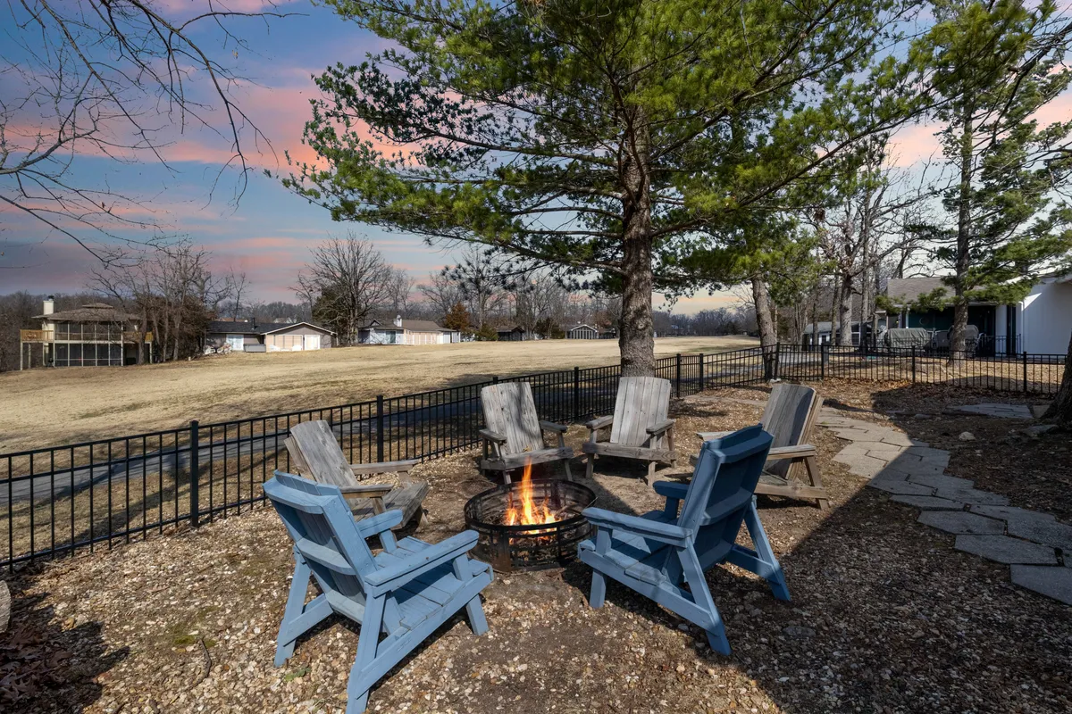 An inviting outdoor scene with a fire pit surrounded by Adirondack chairs and encircled by mature trees. The setting evokes cozy evenings, communal gatherings, and the enjoyment of nature as dusk falls.