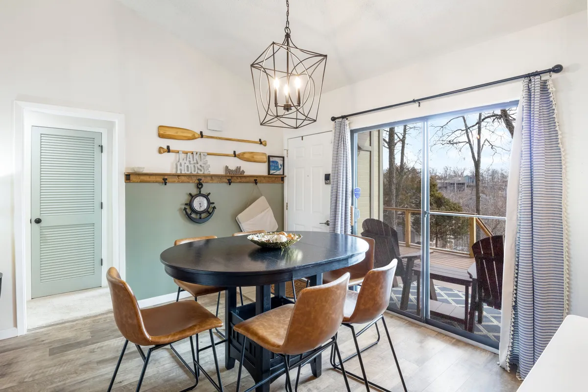 A cozy dining area with a round black table set beneath a hanging light fixture, flanked by wooden paddles on the wall and a large sliding glass door letting in natural light. The setting is perfect for casual gatherings, morning coffee, and celebratory dinners.