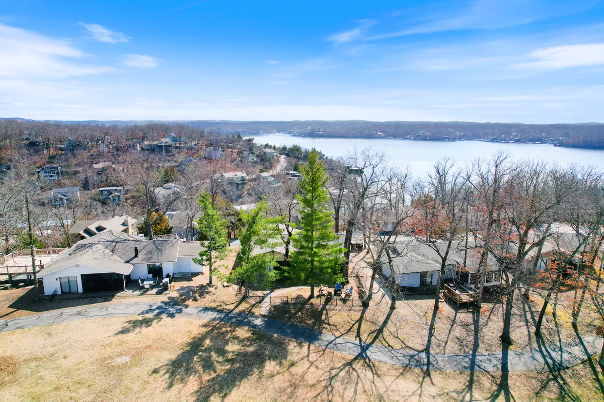 A relaxing exterior shot featuring a lakeside view with green trees, a residential area, and a winding pathway. The scene suggests a peaceful place for boating, picnicking, or simply taking in the natural beauty—a perfect setting for making memories or gathering by a firepit at day’s end.